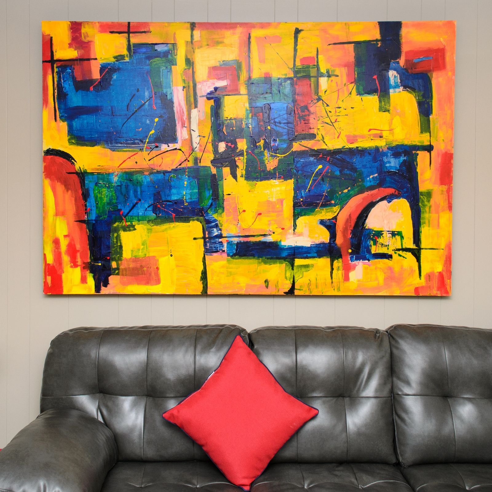 Bright abstract painting above a leather sofa with a red cushion.