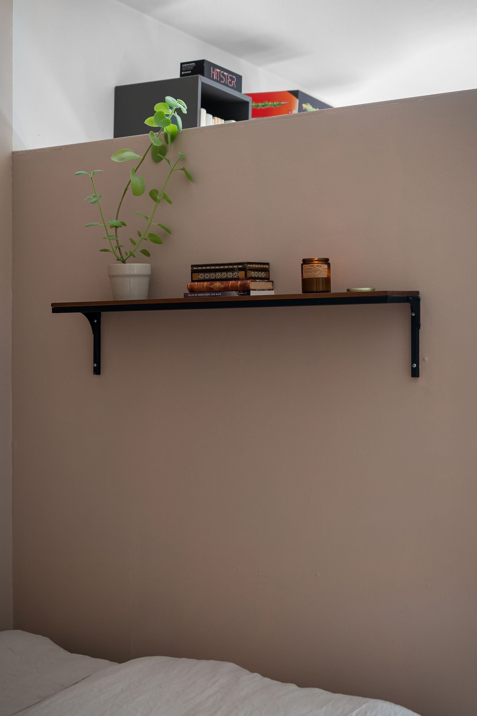 Wall shelf above a bed holding books, a plant and a candle