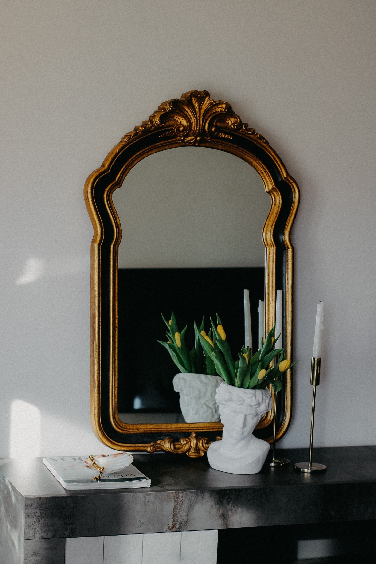 Gold ornate mirror above a mantel with flowers in a bust vase and candles.