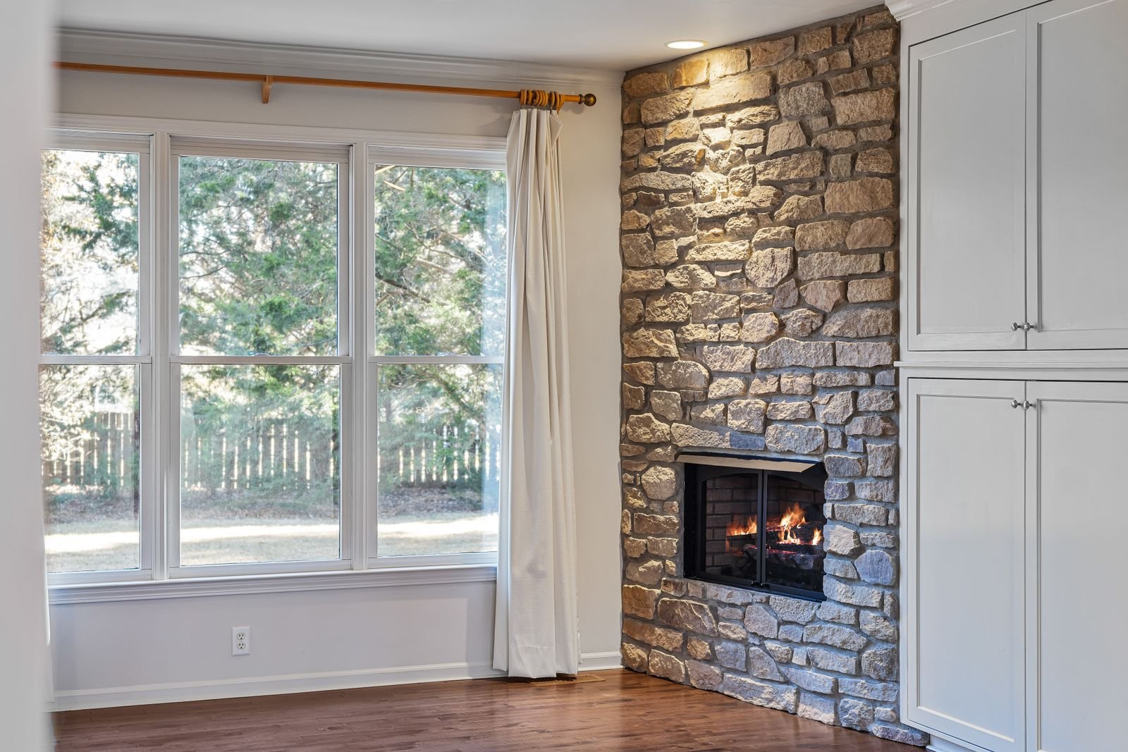 Living room with a stone fireplace, large windows and a hardwood floor.