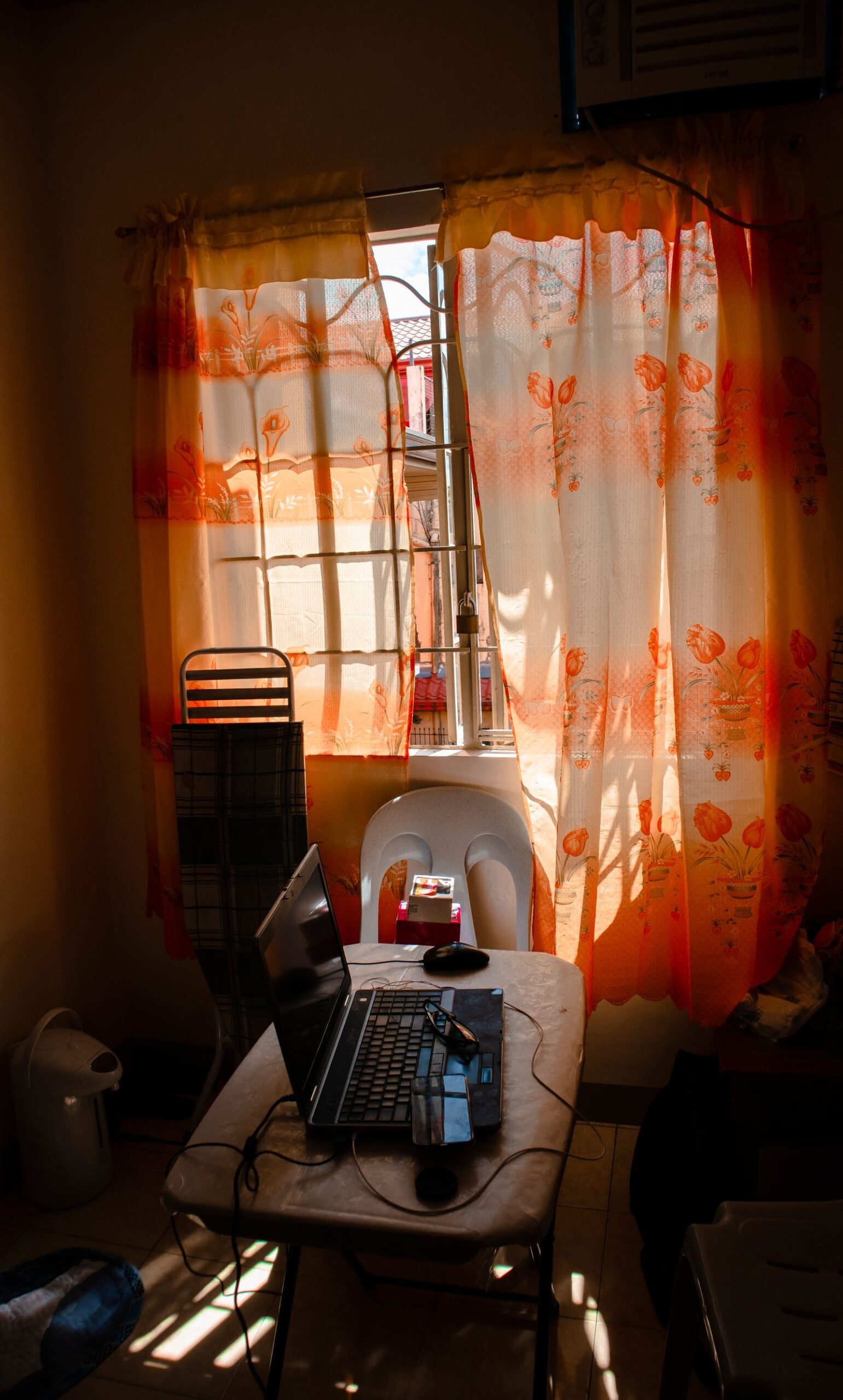 Laptop on a small table in a sunlit room with patterned curtains.