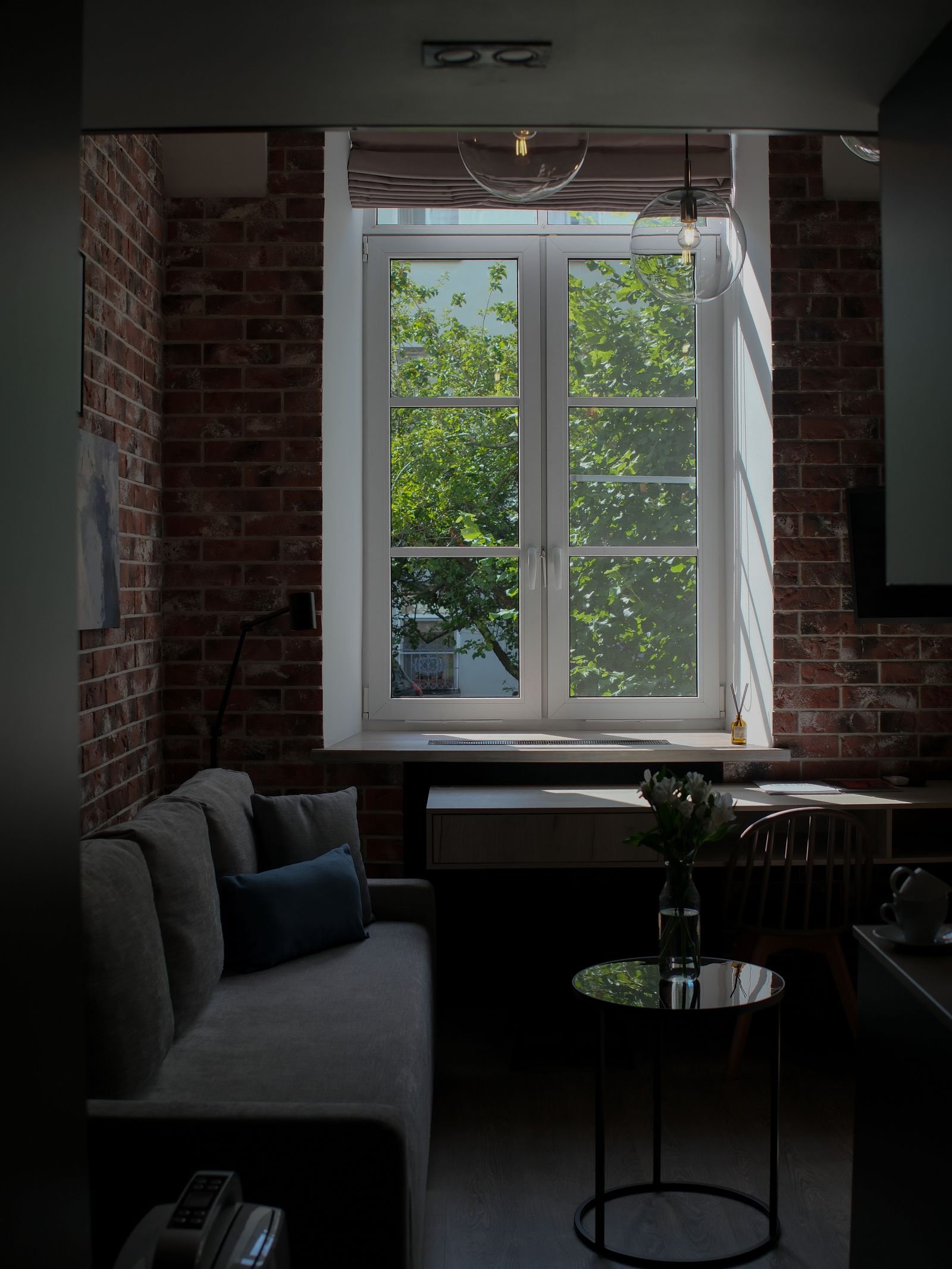 Sunlit living room with a brick wall, sofa and desk by a large window.