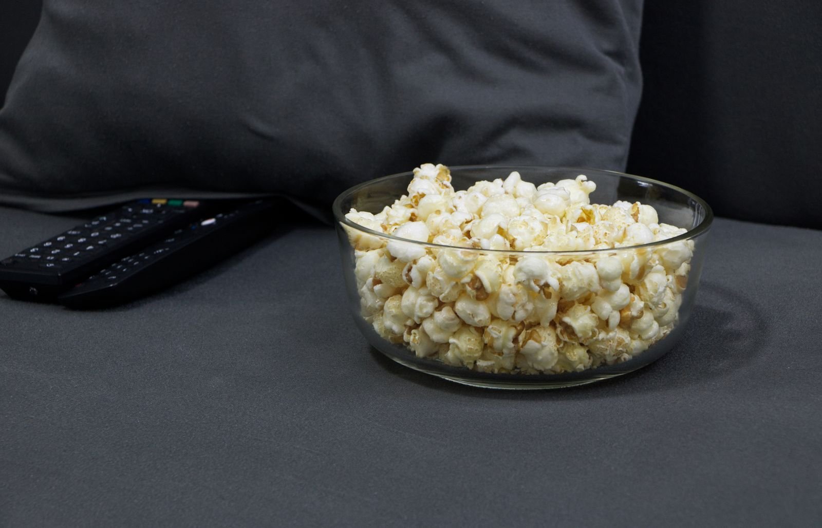 Glass bowl of popcorn beside TV remotes on a gray sofa.
