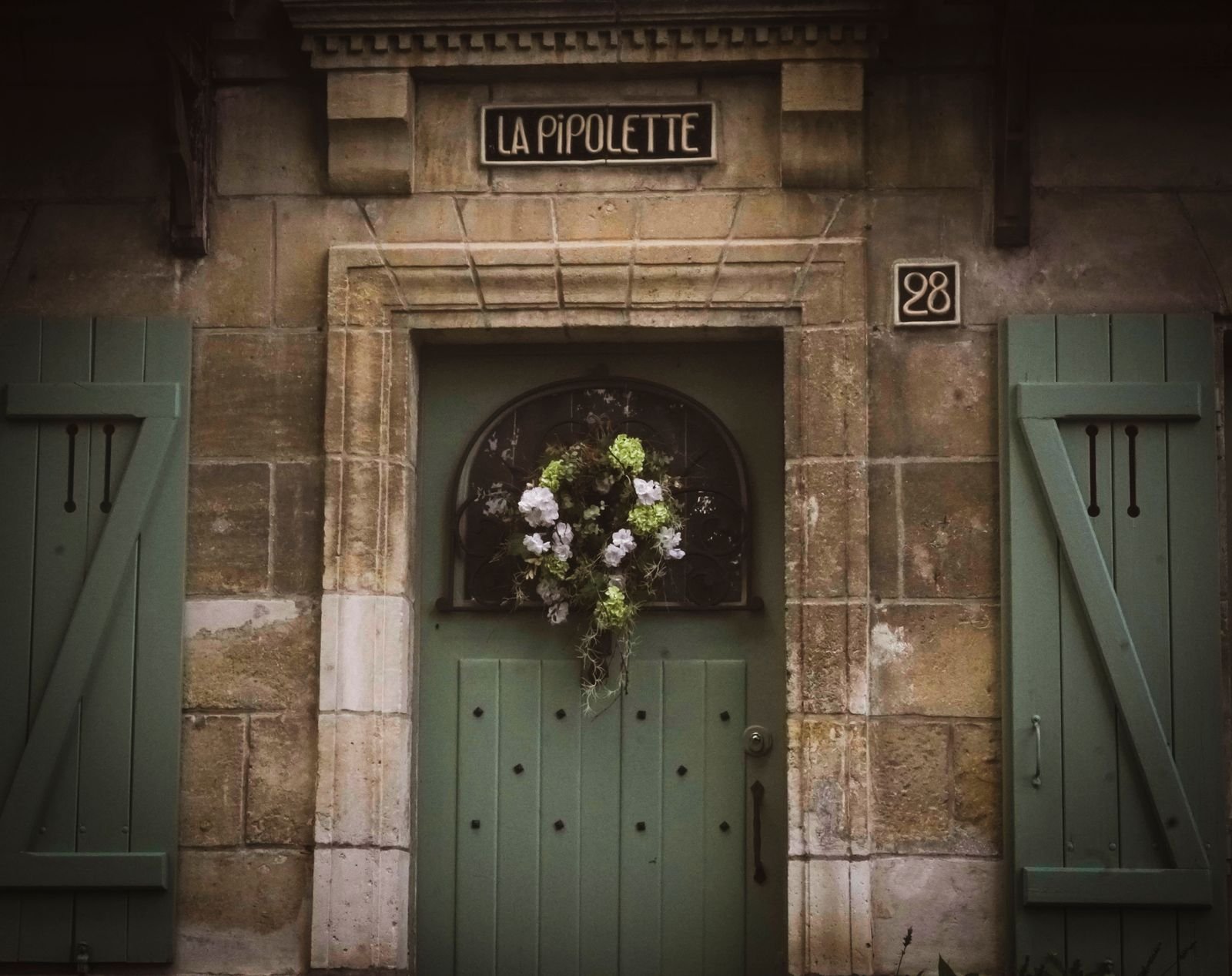 Rustic green front door with a floral wreath and shutters.
