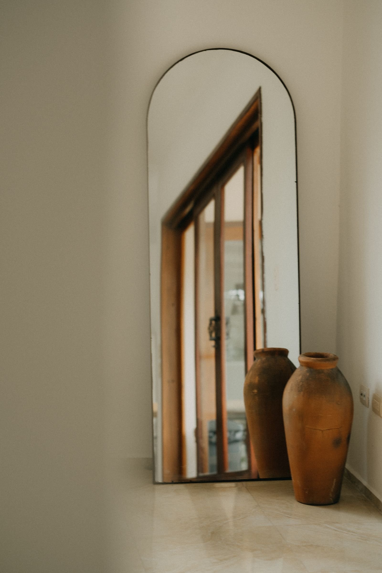 Large arched mirror beside terracotta vases in a warm interior.