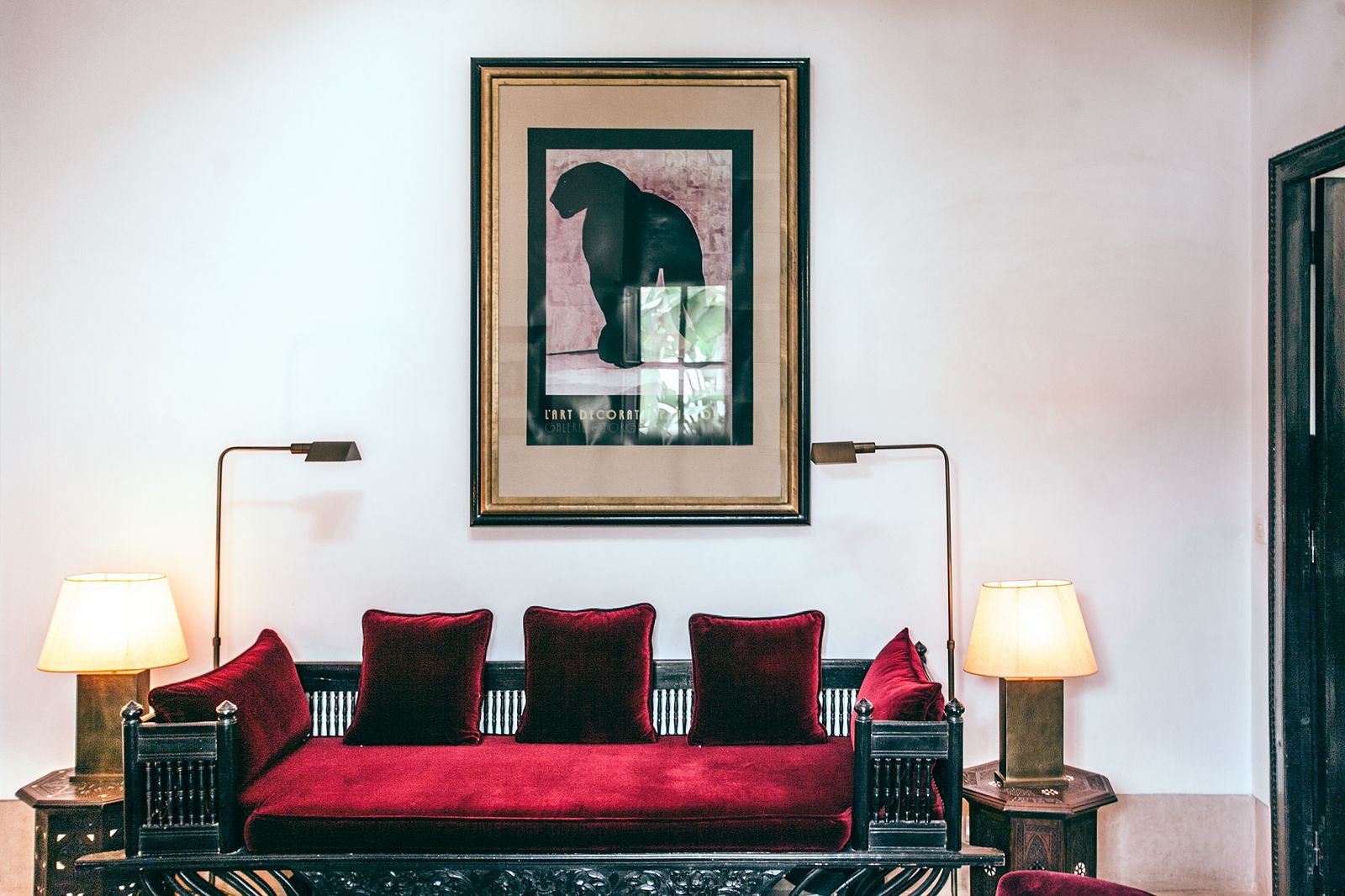 Sofa with red velvet pillows between two lamps below a wall painting.