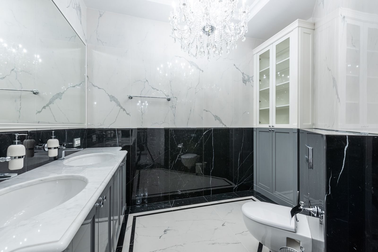 Bathroom with marble walls, white sinks and a glowing chandelier.
