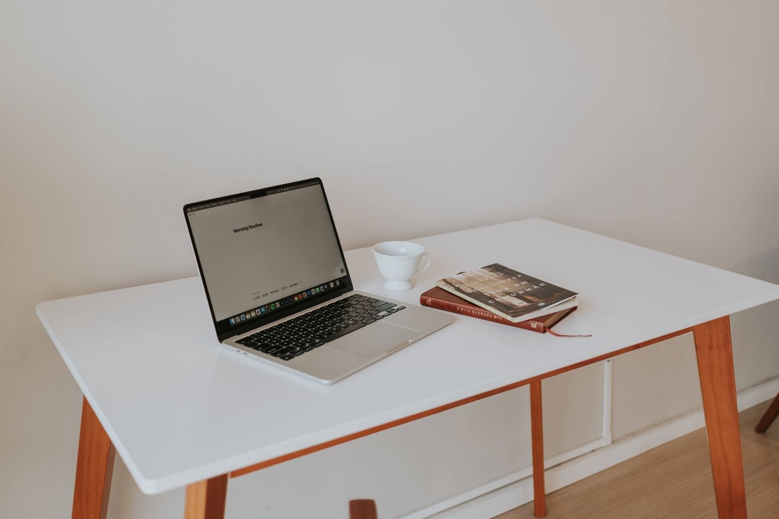 Minimalist white desk with wooden legs, a laptop, a coffee cup and books.