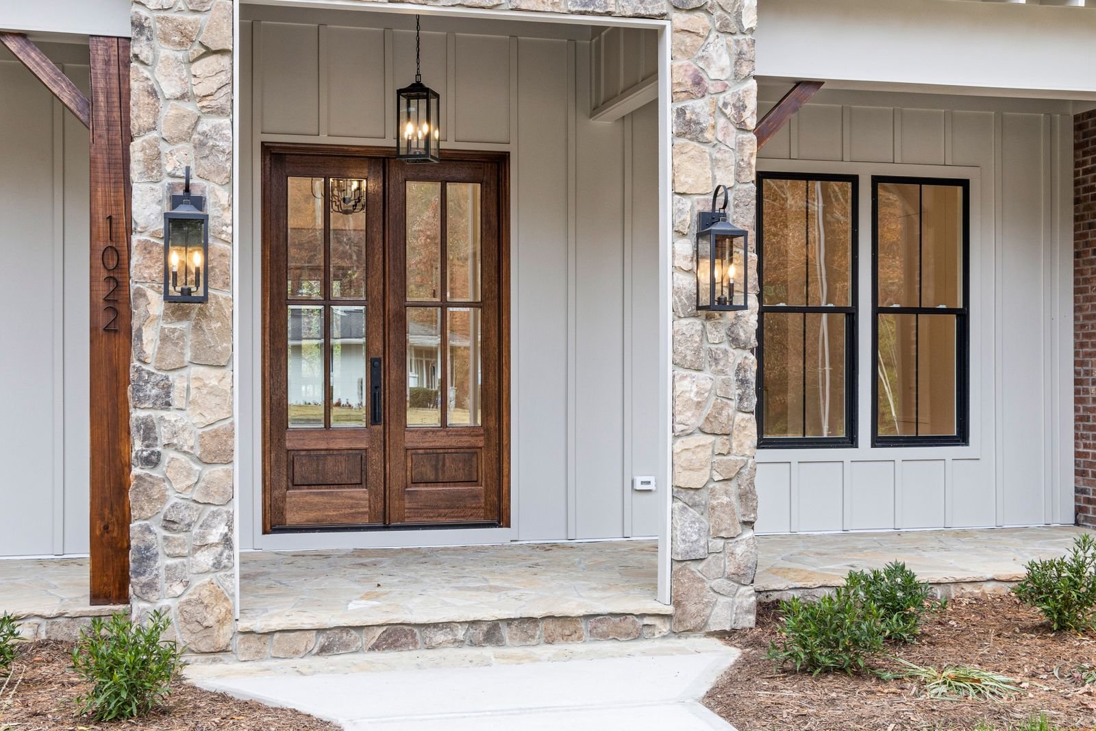 Modern stone house entrance with wooden double doors and lantern-style lights.