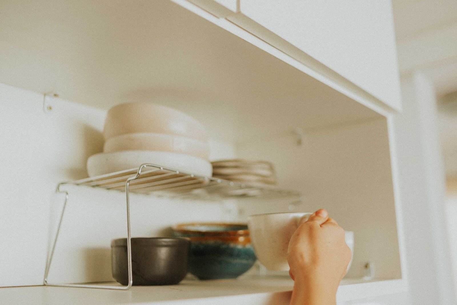 Hand reaching for a mug in a kitchen cabinet with stacked dishes.