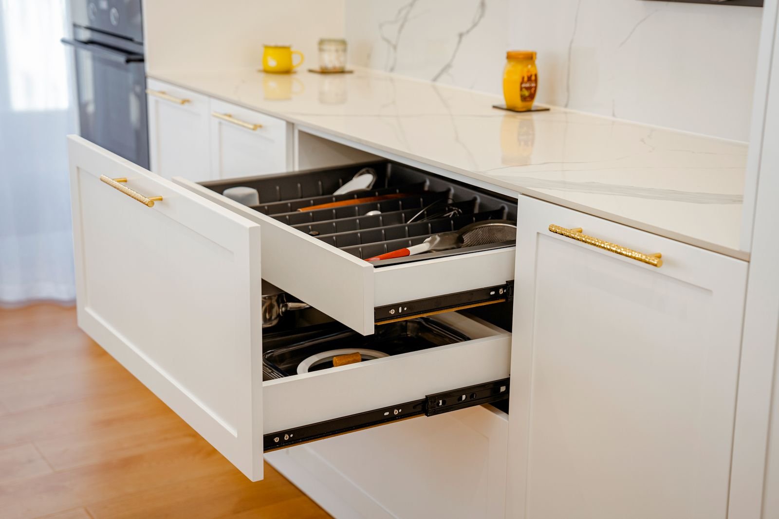 Open white kitchen drawer with utensils beside a marble counter.