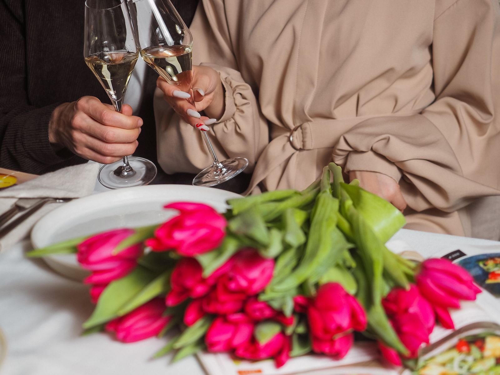 Hands toasting with champagne glasses over pink tulips on a table.