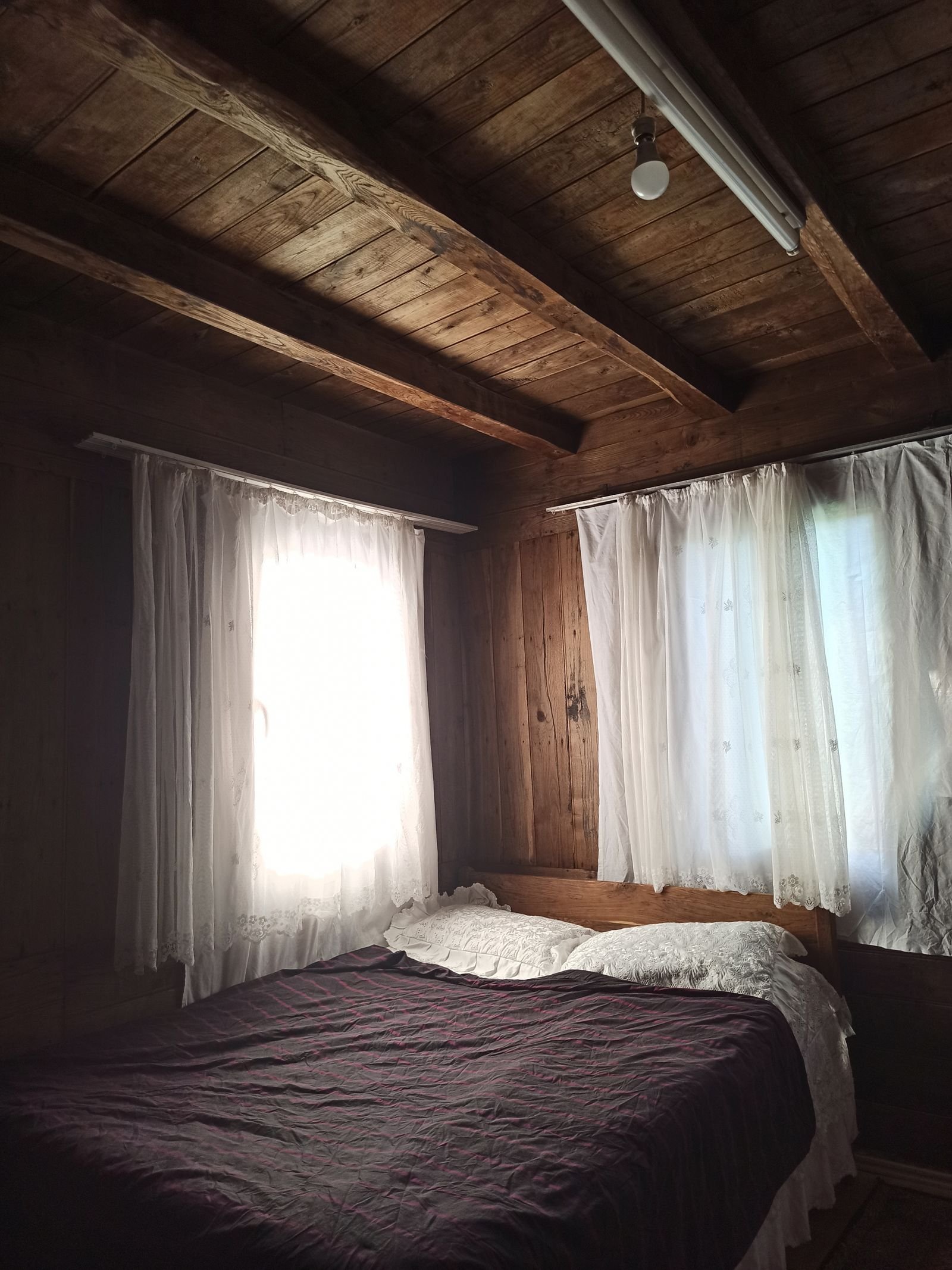 Rustic bedroom with wooden walls, a wooden ceiling and white curtains.