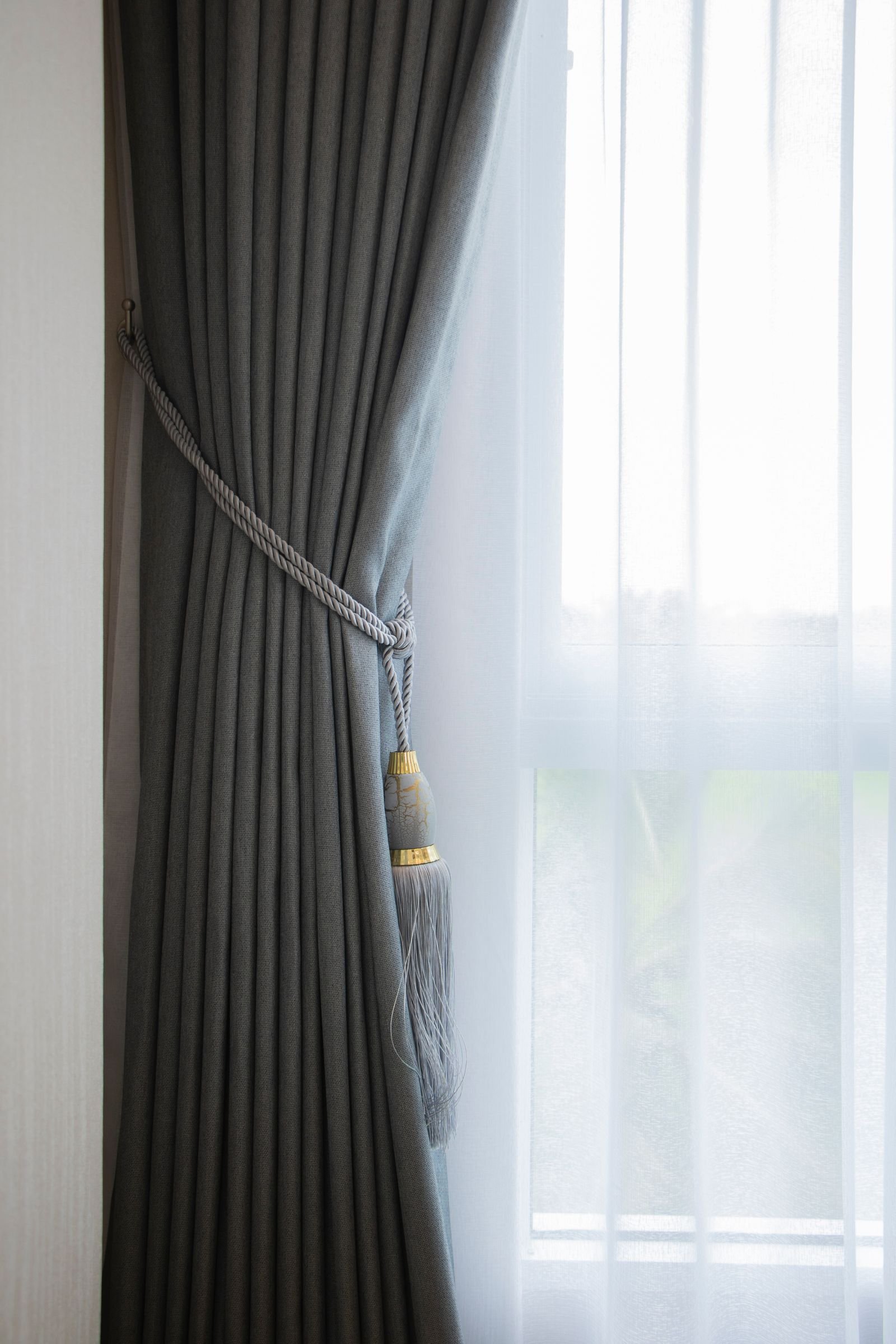 Soft gray curtain held with a tassel tieback over a sheer white curtain